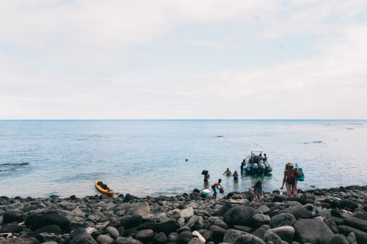People near a rocky shore with a boat and kayak on the water.