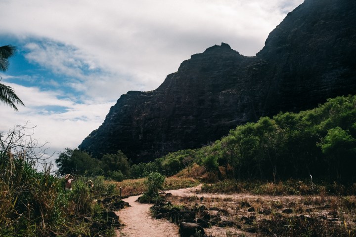 Narrow dirt path in rocky landscape with bushes and large dark cliff.