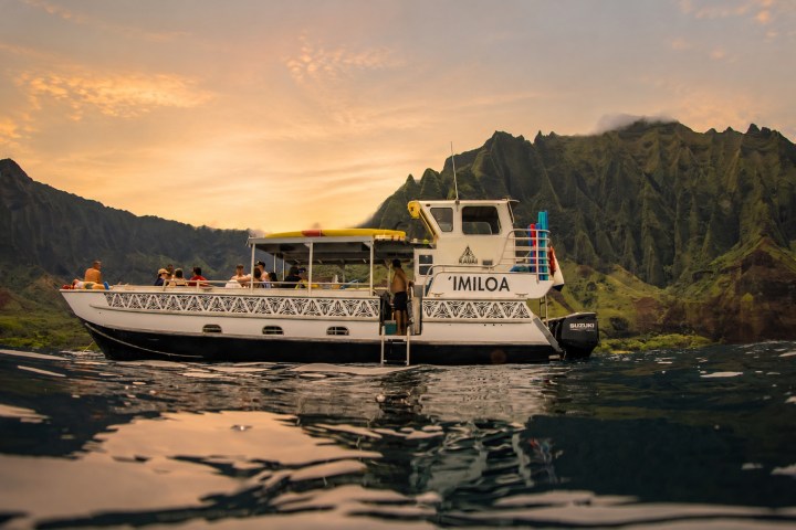 Boat named Imiloa on water with green mountains in background during sunset.