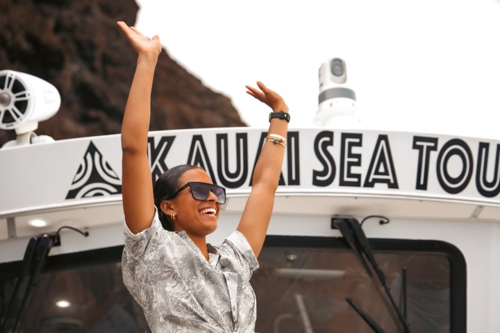 Woman smiling with raised arms in front of a Kauai Sea Tours boat.