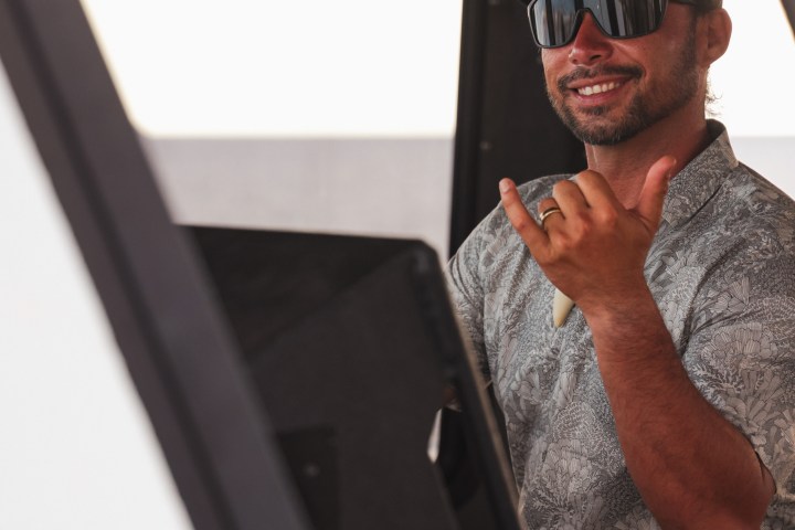 Smiling person with sunglasses and cap, making a hang loose gesture inside a vehicle.