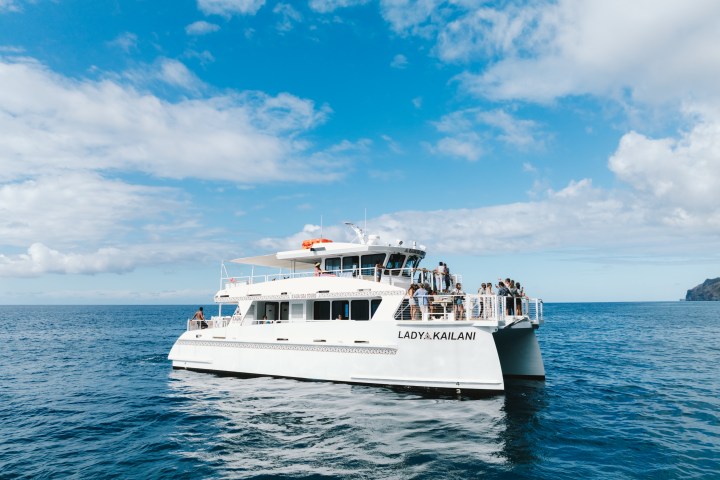 White catamaran with people on deck, sailing on blue ocean under a partly cloudy sky.