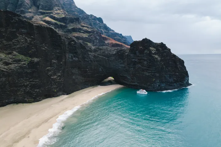 Catamaran near rugged cliffs and sandy beach with turquoise water and cloudy sky.