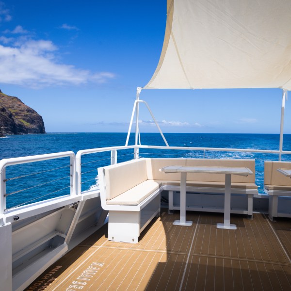 Boat deck with white canopy, seating, tables, ocean view, and cliffs.