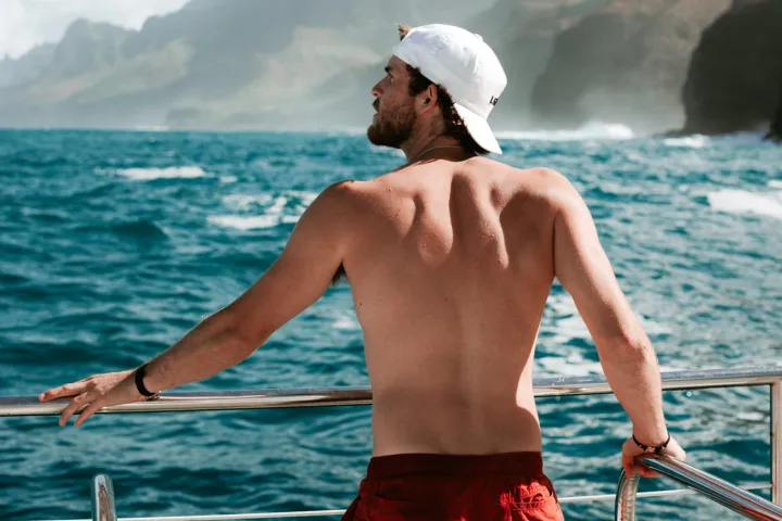 Man in red shorts and cap on boat, overlooking ocean and mountains.