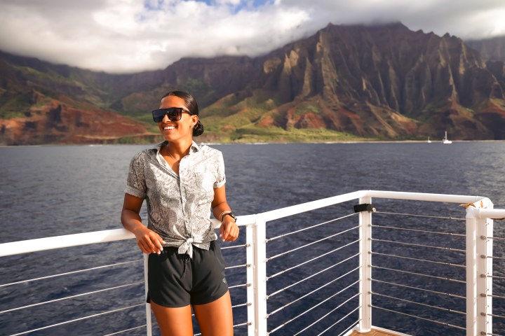 Woman in sunglasses smiles on a boat deck with mountain cliffs and ocean in the background.