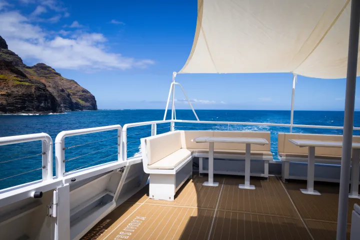 Boat deck with benches and tables, ocean view, and cliffs in the background.