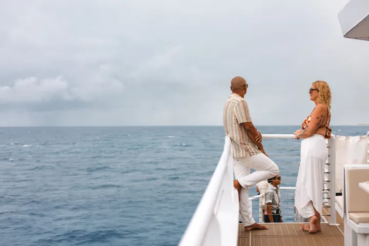 Two people stand on a boat deck, looking at the ocean under a cloudy sky.