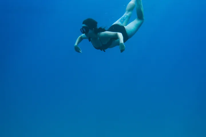 Person snorkeling underwater in clear blue sea with rocky seabed visible.