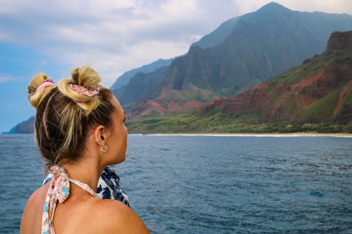 Person on a boat looking at a mountainous coastline under a cloudy sky.