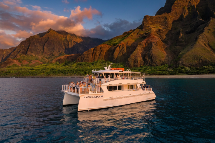 Tour boat on ocean near dramatic mountain cliffs under a sunset sky.