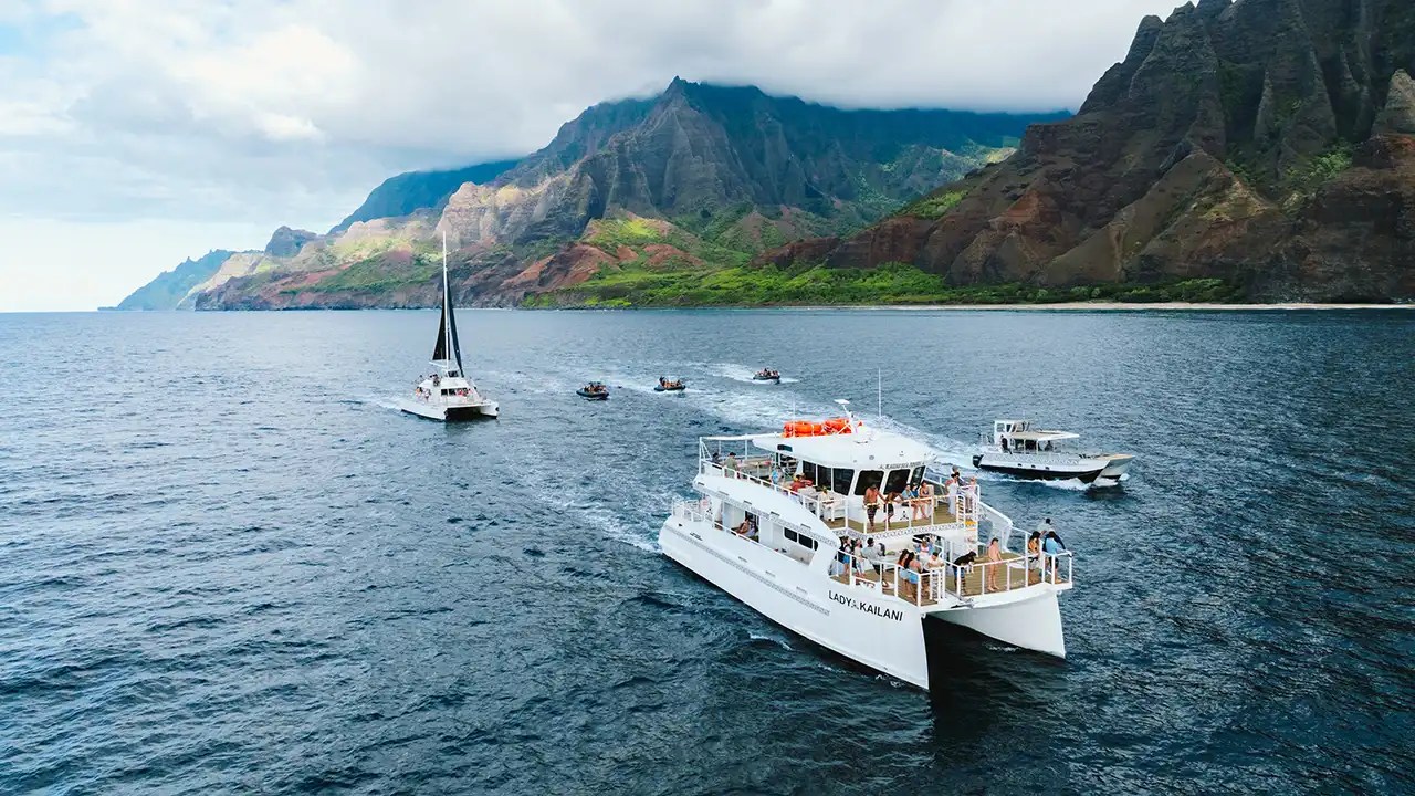 Catamaran and boats sailing near mountainous coastline under partly cloudy sky.