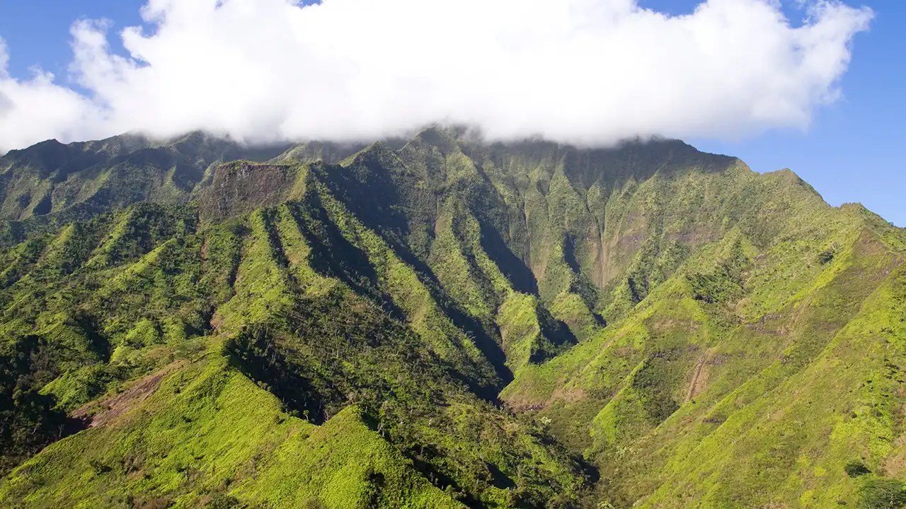 Lush green mountain range with clouds hovering above the peaks under a clear blue sky.