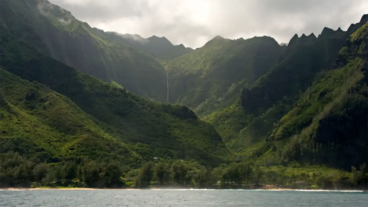 Lush green mountains with a waterfall, viewed from the ocean under a cloudy sky.