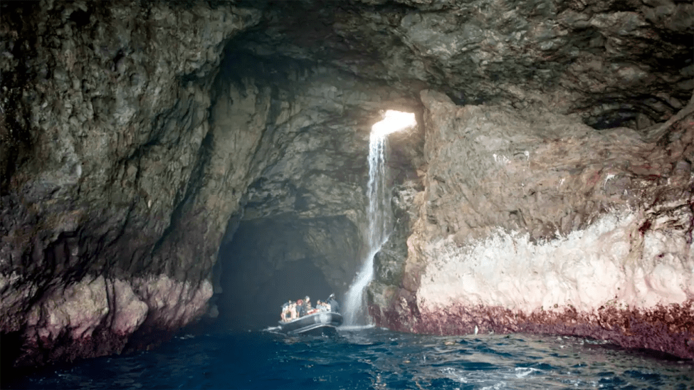 Boat under cave waterfall with rocky walls and blue water.