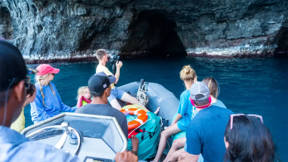 People on a boat approaching a cave entrance on a rocky shore.