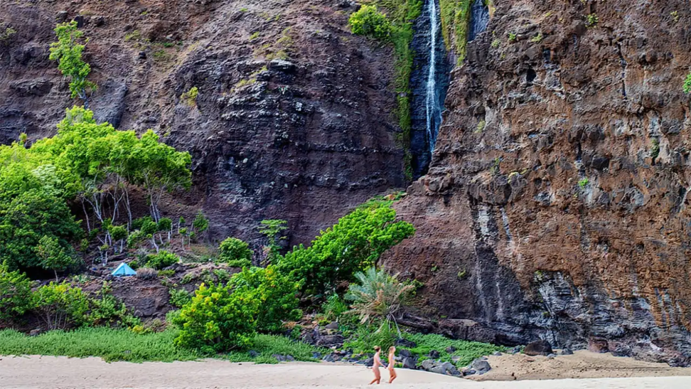 Two people walking on a beach near a cliff with a waterfall.