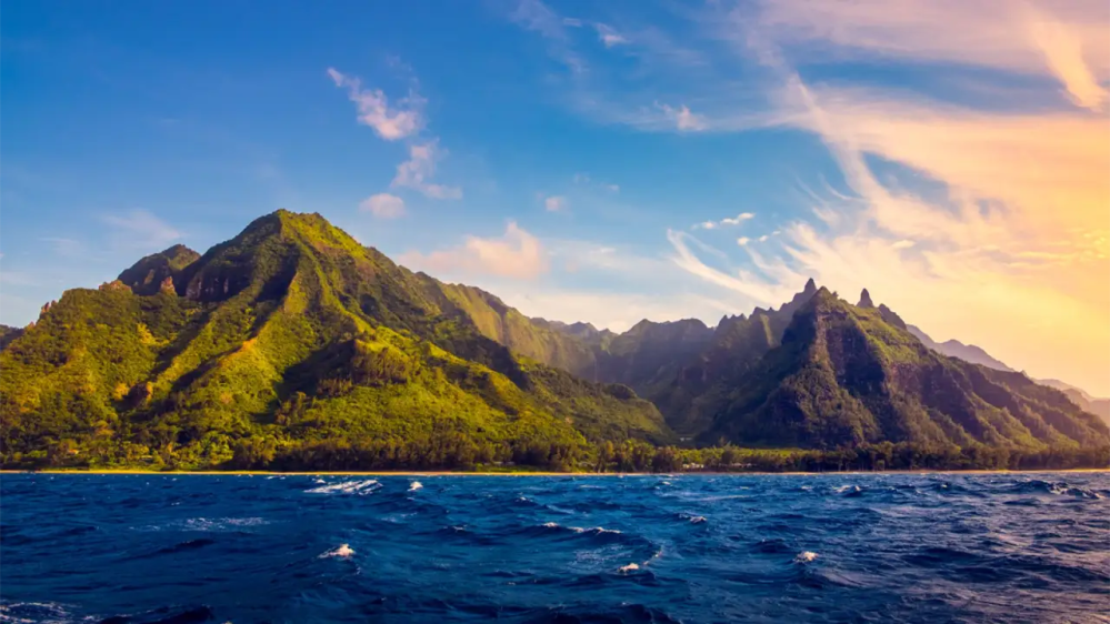 Ocean foreground with lush green mountains under a clear blue sky at sunset.